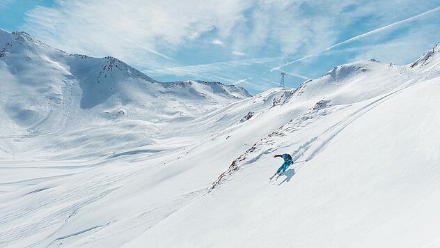Skifahrer in türkisblau auf verschneitem Berghang in weiter Winterlandschaft mit wolkenverziertem Himmel und Alpenkulisse.