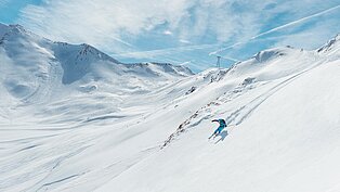 Skifahrer in türkisblau auf verschneitem Berghang in weiter Winterlandschaft mit wolkenverziertem Himmel und Alpenkulisse.