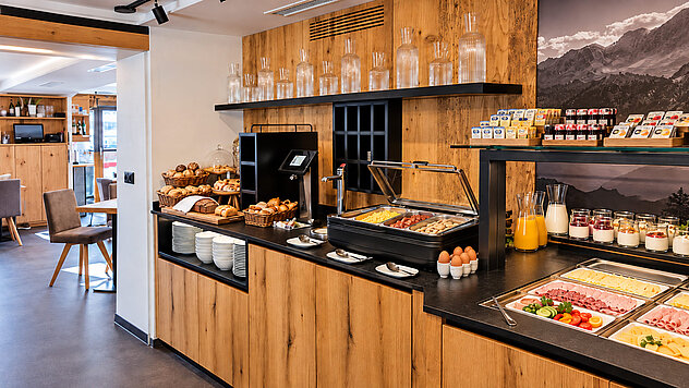 Reichhaltiges Frühstücksbuffet im Valuella mit frischem Brot, Gebäck, Eiern, warmen Speisen, Wurst, Käse, Müsli und Säften an einer modernen Holztheke.