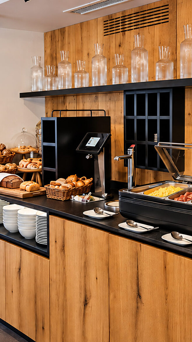 Reichhaltiges Frühstücksbuffet im Valuella mit frischem Brot, Gebäck, Eiern, warmen Speisen, Wurst, Käse, Müsli und Säften an einer modernen Holztheke.