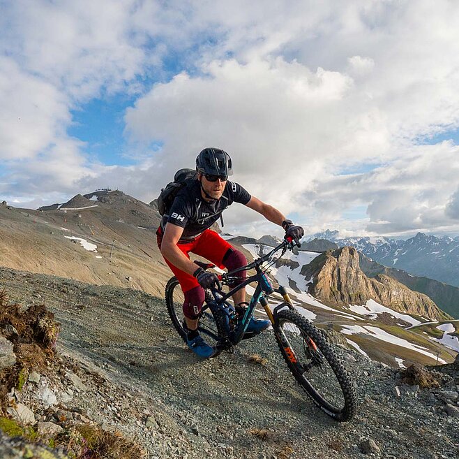 Mountainbiker fährt auf steinigem, alpinem Pfad mit spektakulärer Aussicht über verschneite Gipfel und Täler unter blauem Himmel.