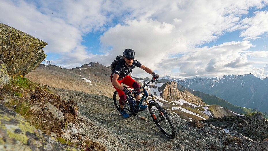 Mountainbiker fährt auf steinigem, alpinem Pfad mit spektakulärer Aussicht über verschneite Gipfel und Täler unter blauem Himmel.