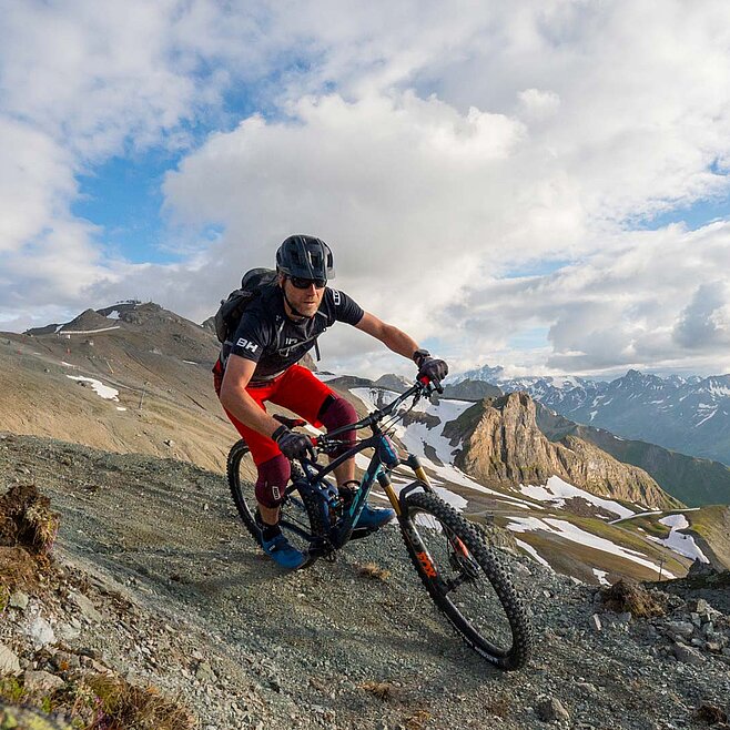 Mountainbiker fährt auf steinigem, alpinem Pfad mit spektakulärer Aussicht über verschneite Gipfel und Täler unter blauem Himmel.