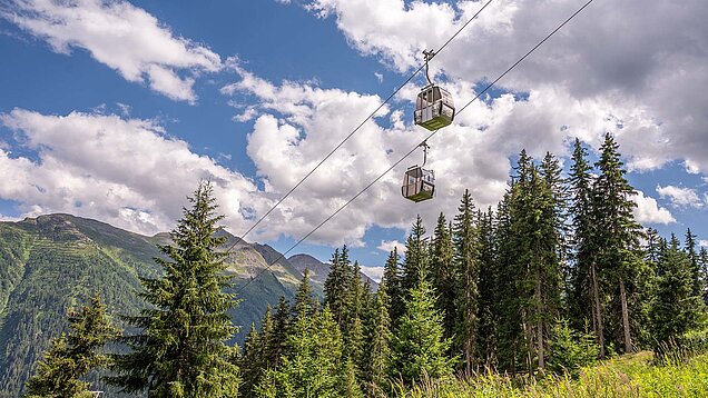 Zwei Gondeln einer Seilbahn schweben über einem dichten Nadelwald, darüber blauer Himmel mit großen Wolken.