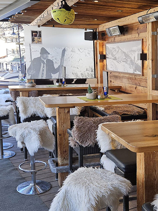 Sonnige Hotelterrasse im alpinen Stil mit Holztischen, Sitzbänken, weichen Fellauflagen und Bergmotiven an den Wänden.