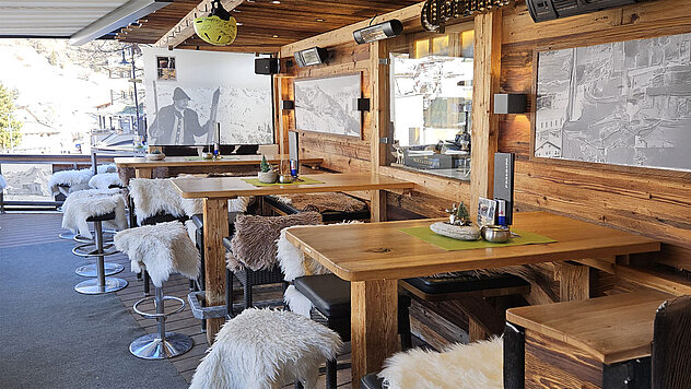 Sonnige Hotelterrasse im alpinen Stil mit Holztischen, Sitzbänken, weichen Fellauflagen und Bergmotiven an den Wänden.