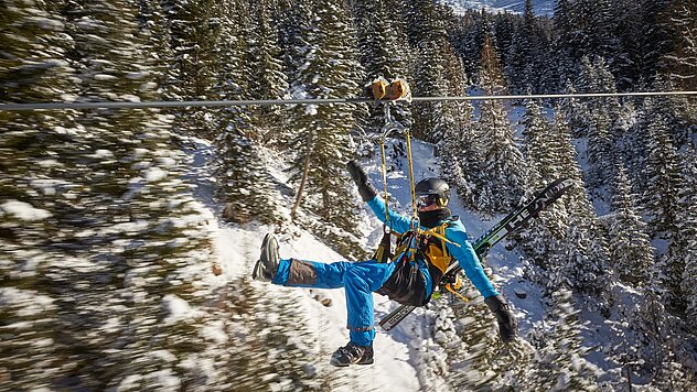 Person beim Ziplining über verschneitem Waldtal, in blauer Ausrüstung mit Helm und Seilgurt, winterliche Bergkulisse im Hintergrund.