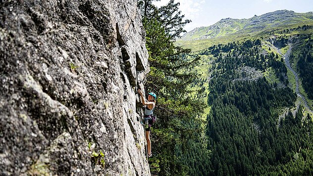 Kletterin am steilen Fels in Ischgl, umgeben von grüner Berglandschaft und alpinem Panorama.