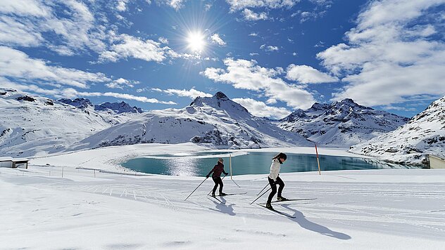 Zwei Personen beim Langlaufen nahe einem türkisblauen Bergsee, umgeben von schneebedeckten Gipfeln unter strahlender Sonne.
