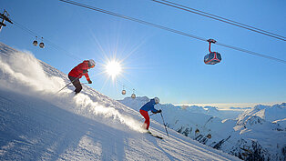 Zwei Skifahrer auf sonniger Abfahrt mit Gondelbahn darüber, Pulverschnee wirbelt auf, Alpenpanorama im Hintergrund.