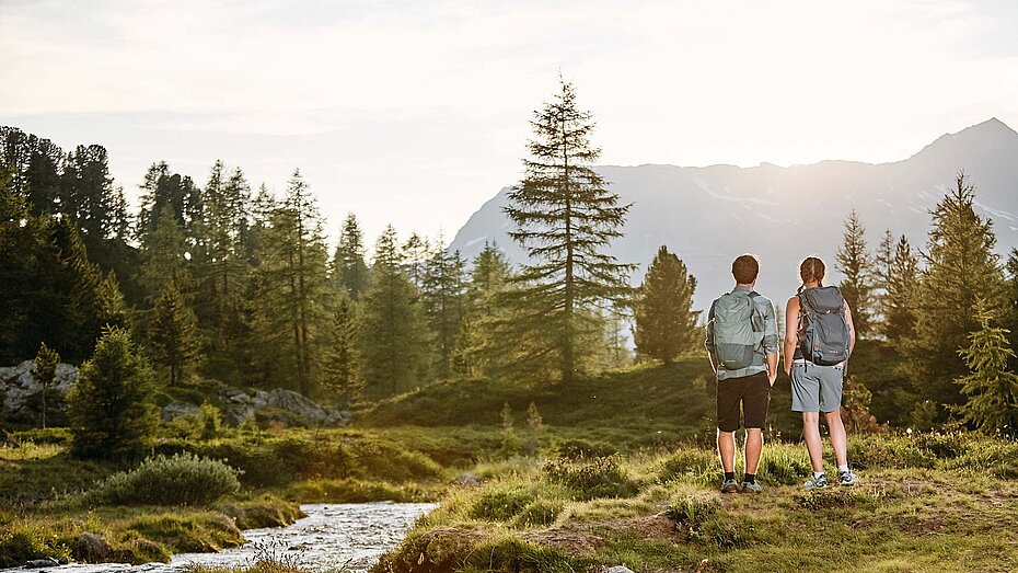 Zwei Wanderer blicken über alpine Landschaft mit Bach, Bäumen und Bergkulisse im sanften Licht der untergehenden Sonne.