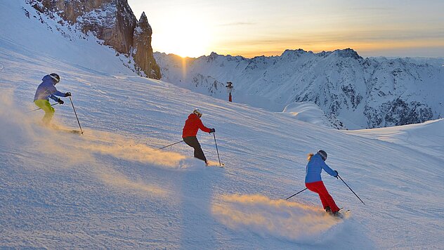 Drei Skifahrer fahren parallel im Pulverschnee bei Sonnenaufgang durch verschneite Berglandschaft mit weitem Ausblick.