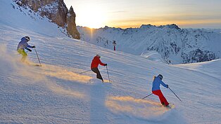 Drei Skifahrer fahren parallel im Pulverschnee bei Sonnenaufgang durch verschneite Berglandschaft mit weitem Ausblick.