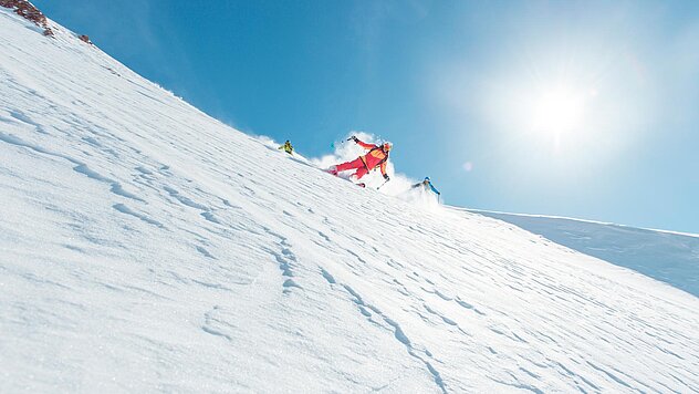 Skifahrer in rotem Outfit fährt bei Sonnenschein eine steile, verschneite Piste mit Schwung und hoher Geschwindigkeit hinab.