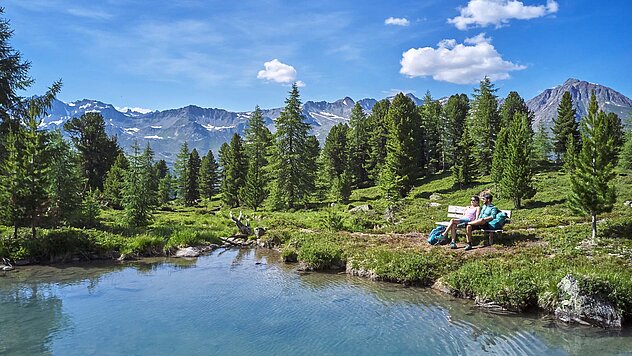 Klarer Gebirgssee mit Wanderern an der Uferwiese, umgeben von Wald, Alpenpanorama und blauem Himmel mit weißen Wolken.