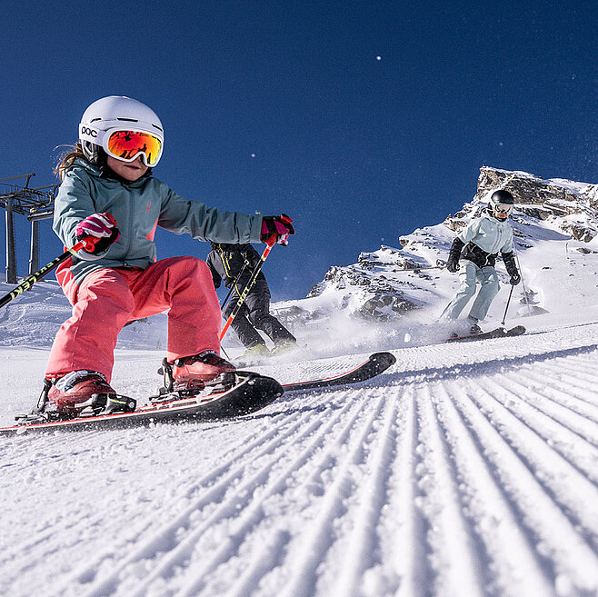 Kind beim Skifahren auf perfekt präparierter Piste in Ischgl, dahinter zwei Erwachsene im Schnee, strahlend blauer Himmel und verschneite Alpenberge.