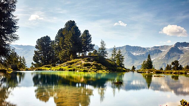 Bergsee mit klarer Spiegelung, bewaldeter Insel, umgeben von Alpenlandschaft und blauem Himmel mit leichten Wolken.