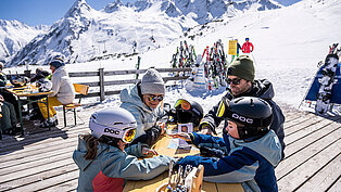 Familie mit zwei Kindern in Skibekleidung auf einer Sonnenterrasse im Skigebiet Ischgl, im Hintergrund verschneite Berge und Skiausrüstung.