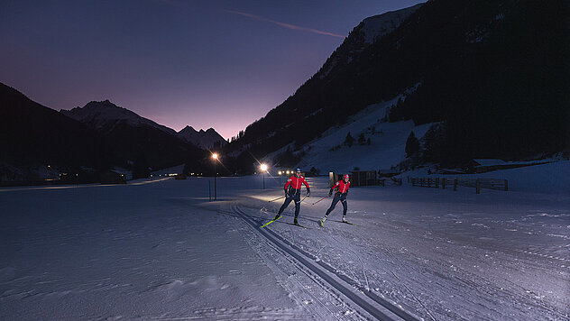 Zwei Personen beim Langlaufen auf beleuchteter Nachtloipe in Ischgl, verschneites Tal und Berge bei Dämmerung.