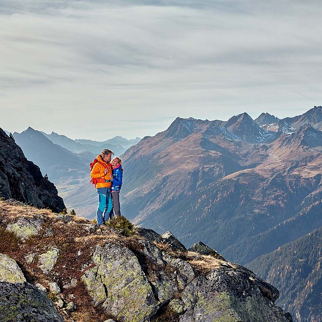 Alleinstehender Wanderer auf Felsvorsprung mit Blick auf schroffe Alpenkette unter bewölktem Himmel – kraftvoll und still.