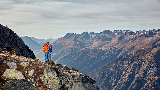 Alleinstehender Wanderer auf Felsvorsprung mit Blick auf schroffe Alpenkette unter bewölktem Himmel – kraftvoll und still.