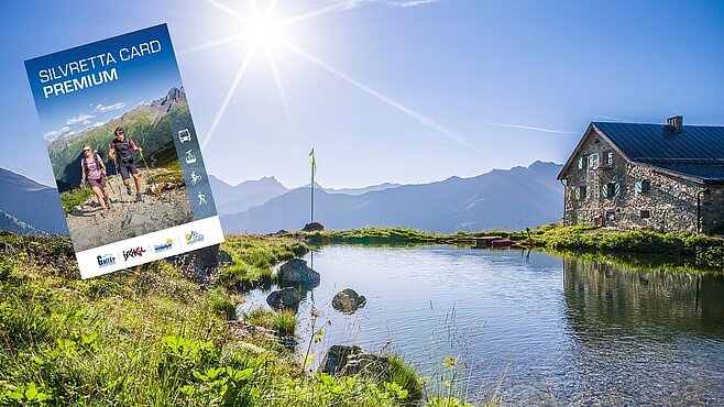 Bergsee mit kleiner Hütte am Ufer und Sonnenschein, im Vordergrund grüne Almwiese; links eingeblendet die Silvretta Card Premium.