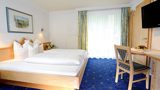 Hotelzimmer mit Doppelbett, Tisch und Stuhl, Fernseher an der Wand und Balkonfenster mit Vorhängen