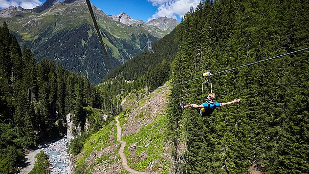 Person beim Ziplining über bewaldetem Tal mit Wasserfall, inmitten steiler Berghänge und beeindruckender Alpenkulisse.