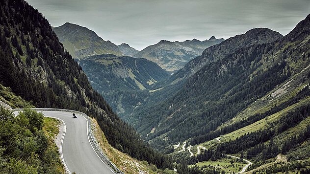 Alpenpassstraße schlängelt sich durch dramatische Berglandschaft mit steilen Hängen, tiefem Tal und Motorradfahrer.