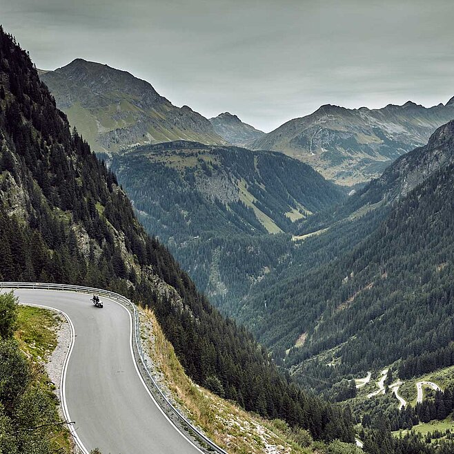 Alpenpassstraße schlängelt sich durch dramatische Berglandschaft mit steilen Hängen, tiefem Tal und Motorradfahrer.