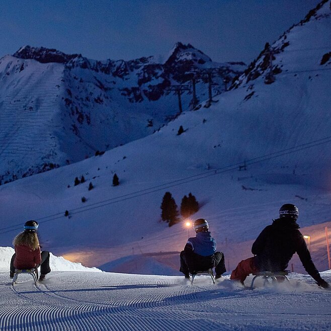 Drei Personen rodeln bei Nacht auf beleuchteter Schneepiste durch verschneite Alpenlandschaft mit markanter Bergkulisse.