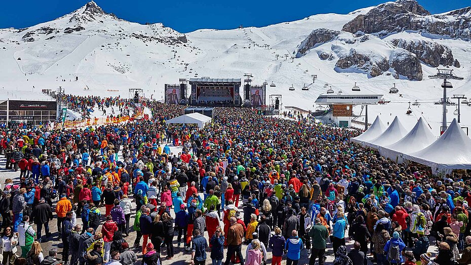 Großes Open-Air-Konzert im Schnee mit bunter Menschenmenge vor Bühne, umgeben von verschneiten Bergen und blauem Himmel.