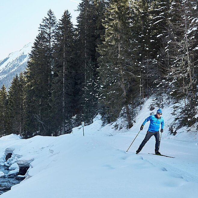 Langläufer auf verschneiter Loipe neben Bachlauf, umgeben von schneebedeckten Bäumen und Berglandschaft im Winterlicht.