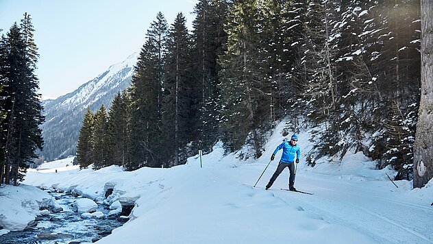 Langläufer auf verschneiter Loipe neben Bachlauf, umgeben von schneebedeckten Bäumen und Berglandschaft im Winterlicht.